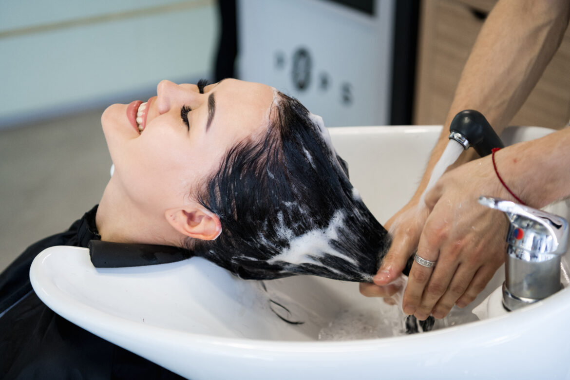 Haircare procedure in beauty salon. Hairdresser is brushing woman’s hair spreading a treatment mask or conditioner