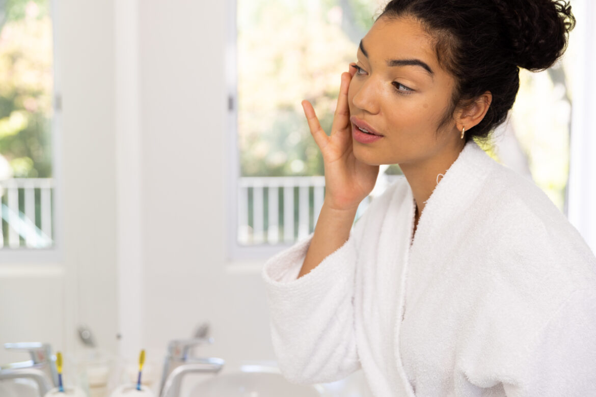 Biracial woman in bathrobe looking in mirror in sunny bathroom. Lifestyle, self care and domestic life, unaltered.