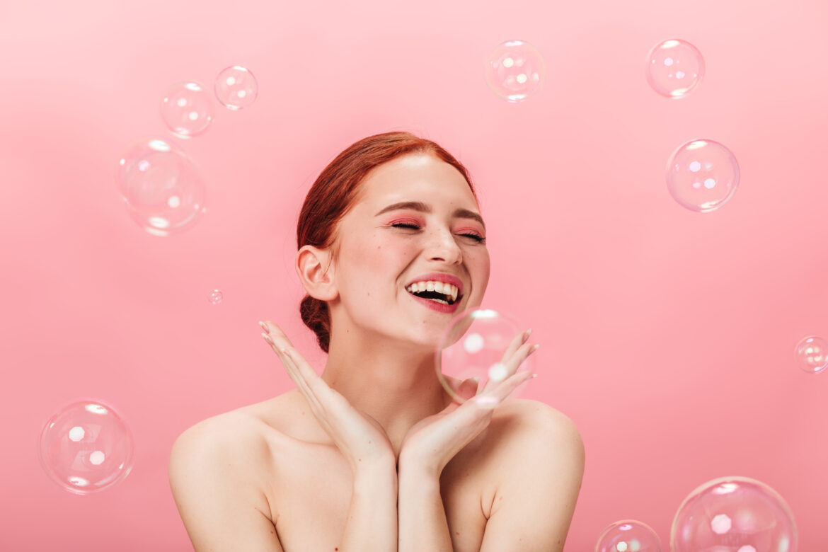 Inspired nude girl posing with soap bubbles. Studio shot of laughing young woman with ginger hair.