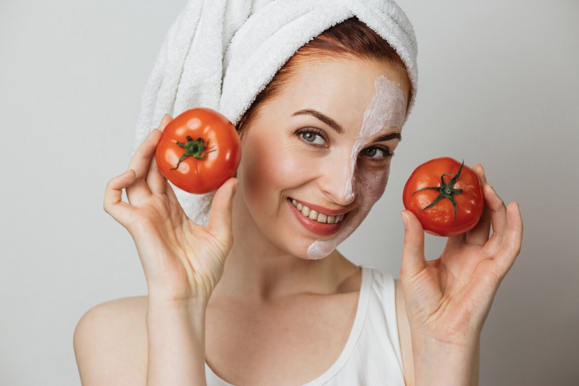 Woman in towel with cosmetic moisturizing mask on half of her face holding two fresh tomato.
